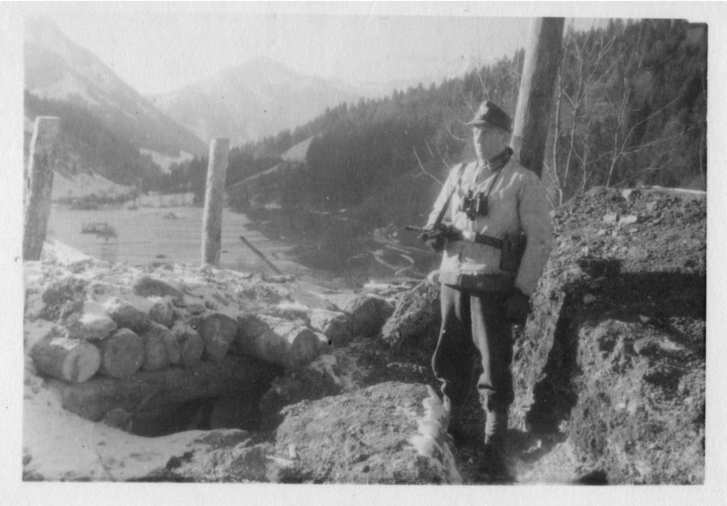 My grandfather during war time in front of a trench. He wears a soldiers shield cap, a light-colored jacket, dark trousers and boots. He is armed with a machine gun and carries binoculars. In the background a wooded mountain range and houses and streets in the valley