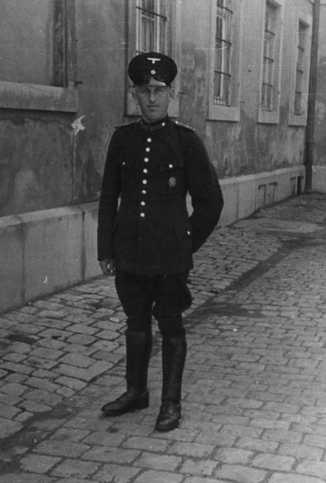 My grandfather in a black police uniform with shield cap and flashing metal buttons. He wears a dark uniform pants with leather boots over them up to the knee.